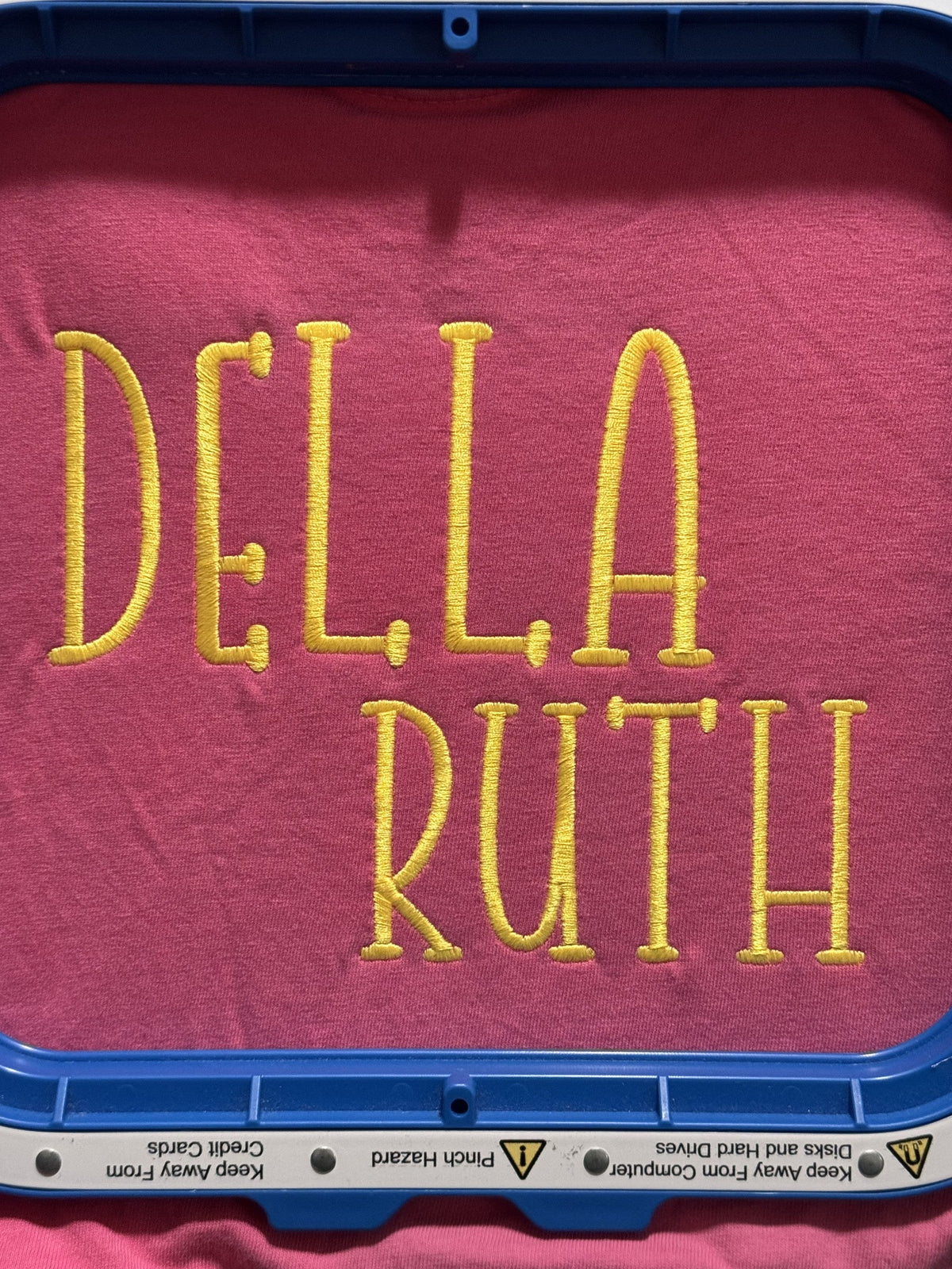 Pink fabric embroidered with the stacked name Della Ruth in yellow thread