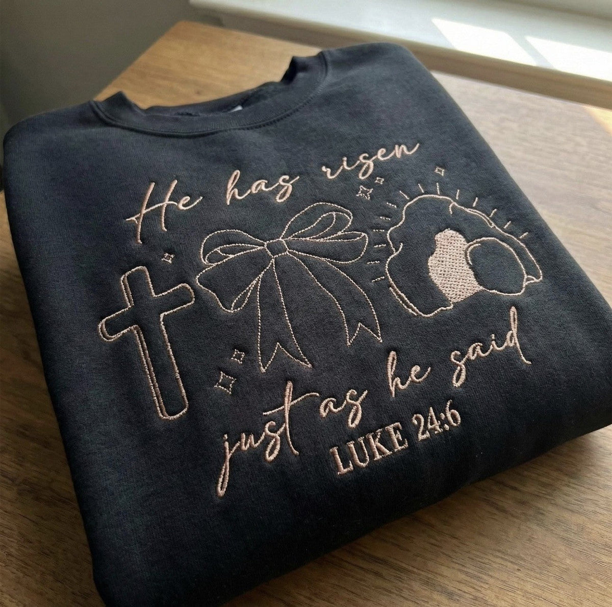 Dark sweatshirt with He Has Risen faith embroidery featuring a cross and leaf motif