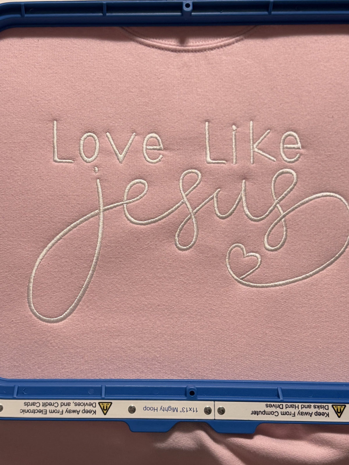 Pink fabric embroidered with the phrase Love Like Jesus shown in an embroidery hoop